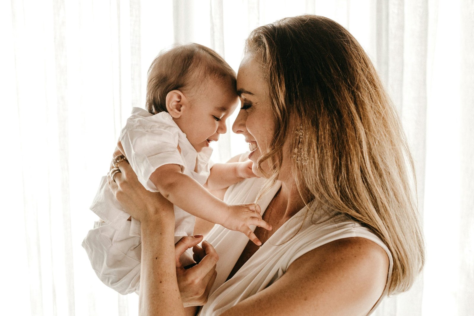 A parent and baby, faces close together, backlit by soft window light